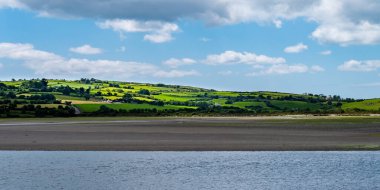 White clouds in the sky, hilly Irish countryside. Green hills on a nice summer day. Irish countryside, County Cork. Green grass field near body of water under white clouds and blue sky