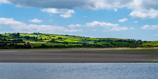 White clouds in the sky, hilly Irish countryside. Green hills on a nice summer day. Irish countryside, County Cork. Green grass field near body of water under white clouds and blue sky