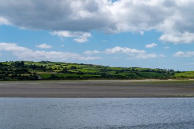 White clouds in the sky, hill, Irish countryside. Green hills on a nice summer day. Irish countryside, County Cork. Green grass field near body of water under white clouds and blue sky