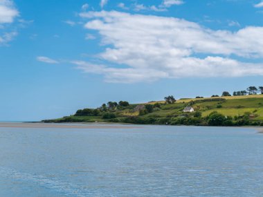 Beautiful sky over a water surface on a summer day. The picturesque green coast of Ireland. Several buildings on the hill. Seascape. Green trees on island