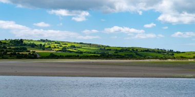 White clouds in the sky, Irish countryside. Green hills on a nice summer day. Irish countryside, County Cork. Green grass field near body of water under white clouds and blue sky