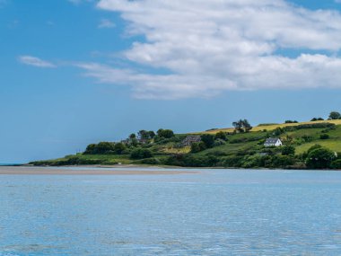 Beautiful sky over a water on a summer day. The picturesque green coast of Ireland. Several buildings on the hill. Seascape. Green trees on island