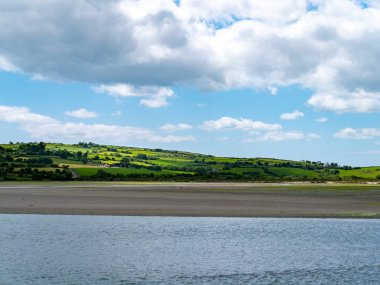 White clouds in the sky, countryside. Green hills on a nice summer day. Irish countryside, County Cork. Green grass field near body of water under white clouds and blue sky