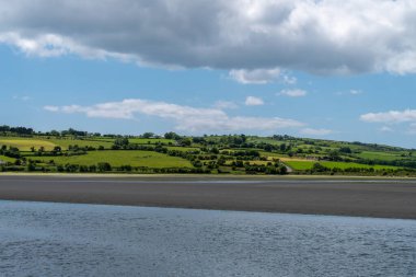 Gökyüzünde beyaz bulutlar, kırsal kesimde. Yazın yeşil tepeler. İrlanda kırsalı, County Cork. Suyun yanındaki yeşil alan, beyaz bulutların ve mavi gökyüzünün altında.