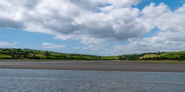 İrlanda kırsalının tepesinde gökyüzünde beyaz kümülüs bulutları. Yeşil tepeler güzel bir yaz gününde. İrlanda kırsalı, County Cork. Yeşil çimen tarlası, su