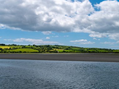 İrlanda kırsalının tepesinde gökyüzünde beyaz kümülüs bulutları. Yeşil tepeler güzel bir yaz gününde. İrlanda kırsalı, County Cork. Beyaz bulutların altında, suyun yanında yeşil çimen tarlası