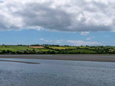 İrlanda kırsalının tepesinde gökyüzünde beyaz kümülüs bulutları. Yeşil tepeler güzel bir yaz gününde. İrlanda kırsalı, County Cork. Suyun yanında yeşil çimen tarlası, beyaz bulutlar