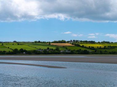 İrlanda kırsalının tepesinde gökyüzünde beyaz kümülüs bulutları. Yeşil tepeler güzel bir yaz gününde. İrlanda kırsalı, County Cork. Suyun yanında yeşil çimen tarlası, bulutlar.