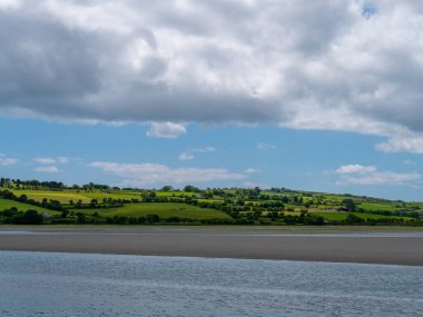 İrlanda kırsalının tepesinde gökyüzünde beyaz kümülüs bulutları. Yeşil tepeler güzel bir yaz gününde. İrlanda kırsalı, County Cork. Suyun yakınında yeşil çimen tarlası