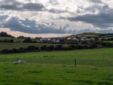 Bulutlu bir gökyüzünün altındaki yeşil tarlaların ve ağaçların arasında bir İrlanda köyü, County Cork. Yazın kırsal İrlanda manzarası, Avrupa köyü. Bulutlu gökyüzünün altındaki yeşil çimen tarlası