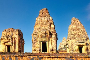 Yüksek çözünürlüklü görüntü, Pre Rup tapınağının üç anıtsal tuğla kulesini yakalıyor, bulutsuz mavi gökyüzünün altında gururla duruyor, Khmer mimari tarzını yansıtıyor..