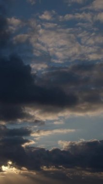 Güneşin ışınları açık mavi gökyüzündeki kalın bulutları delip geçiyor. Arka planda hızla hareket eden bulutlar, video zaman dilimi. Dikey video.