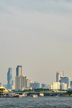 Önünde nehir olan bir şehir silueti. Ufuk çizgisi yüksek binalardan oluşur. Nehirde kayıklar ve arka planda bir köprü var. Gökyüzü puslu. Bangkok