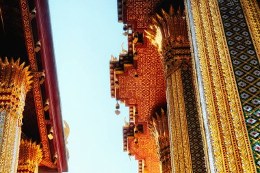 Bu resim Tayland 'daki bir tapınağı betimliyor. Karmaşık altın ve kırmızı desenlerle süslenmiş. Küçük heykeller çatıyı süslüyor. Alçaktan çekilen fotoğraf, arka planda gökyüzünü gösteriyor..