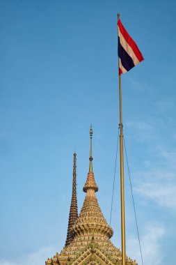 Mavi gökyüzüne karşı bir Tayland tapınağı kulesi ve bayrağı. Tapınak kulesi süslenmiş ve altın rengi var. Bayrak kırmızı, beyaz ve mavi ve bayrak direğinde dalgalanıyor. Gökyüzü açık mavi.