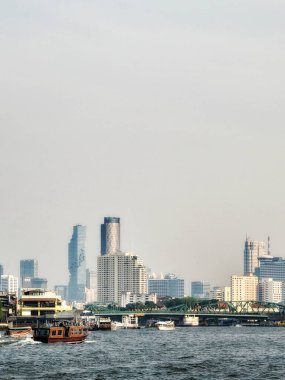 Önünde nehir olan bir şehir silueti. Ufuk çizgisi yüksek binalardan oluşur. Nehirde kayıklar ve arka planda bir köprü var. Gökyüzü puslu ve renkler sessiz. Bangkok