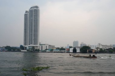 Bangkok, Tayland, 28 Aralık 2018. Kırmızı gölgelikli bir teknenin yelken açtığı dalgalı bir nehrin yanında uzun, beyaz bir bina duruyor. Yukarıda bulutlu gökyüzü beliriyor.