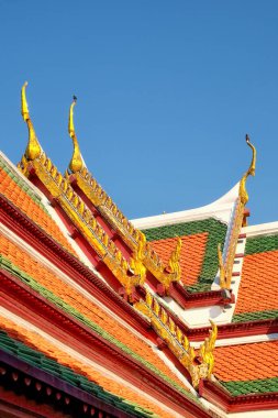 Tayland 'da bir tapınağın çatısında. Çatı, altın süslemelerle süslenmiş birden fazla kırmızı ve yeşil fayans katmanından oluşur. Gökyüzü açık mavi. Tayland mimarisi.