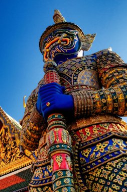 Mavi yüzlü, yeşil ve altın başlıklı, büyük bir kılıç tutan bir dev heykeli, açık mavi bir gökyüzünün altındaki süslü Tayland tapınağının önünde nöbet tutuyor..