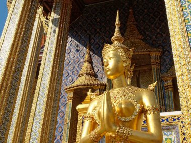 Mavi ve altın renkli mozaik fayanslarla bezenmiş bir tapınağın önünde, geleneksel Tayland kıyafetleri içinde görkemli altın bir kadın heykeli duruyor..