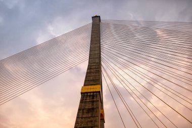 Bulutlu gökyüzünün arka planında, kablo destekli köprünün devasa beton direği görkemli bir şekilde yükselir, kablolar arp gibi dizilmiştir..