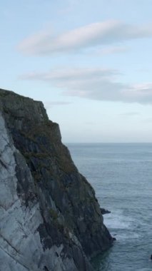 Dik kayalık bir uçurumun tepesinde gururla tünemiş beyaz bir deniz feneri aşağıdaki geniş, safir okyanusu denetliyor. Gökyüzü denizin rengini yansıtıyor, bir tutam kabarık bulutla. Dikey video, yan.