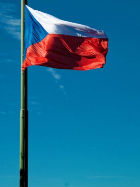 Çek Cumhuriyeti bayrağı rüzgarda dalgalanıyor. Arkasında açık mavi bir gökyüzü olan bayrak direğinde..