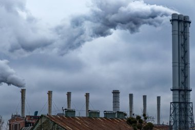 Birden fazla bacası olan bir fabrika gökyüzüne duman yayıyor. Gökyüzü bulutlu ve fabrika arka planda. Duman bacaları uzun ve gri. Birden fazla bacası olan fabrika..