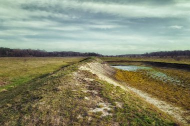 Bir manzara fotoğrafı, ufka doğru uzanan küçük bir göleti ve kirli bir yolu olan çimenli bir tarlayı yakalar. Kuru bir gölün dibi. Sığ bir su kütlesi..