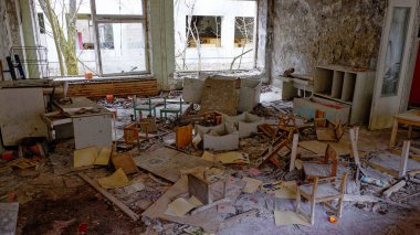 Görüntü dağınık mobilya, enkaz ve duvarların soyulduğu harap bir odayı gösteriyor. Pencereler kirli ve kırık..