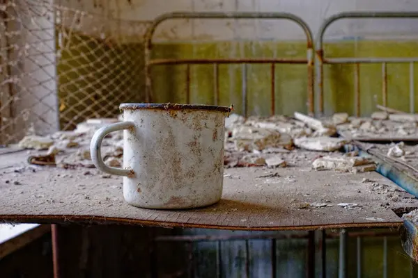 A rusted mug on a dilapidated table in an abandoned room. A white metal mug on a bed in an abandoned building in Chernobyl.