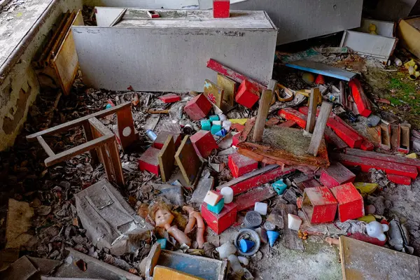 A cluttered room with scattered toys, wooden blocks, a doll, and debris on the floor. The area looks abandoned and in disarray. Scattered furniture and toys in an abandoned kindergarten in Pripyat.