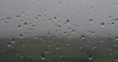 Pencereye konan yağmur damlalarının renkli bir videosu, arka planda yeşil bir alan ve ağaçlar var. pano