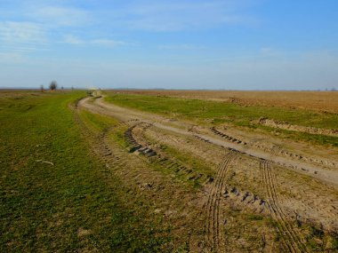 Manzara, mavi gökyüzünün altındaki yeşil tarlaların arasında asfaltsız bir yola sahiptir..