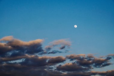 Ay, akşam gökyüzünde dağınık bulutların arasında parlak bir şekilde parlıyor..