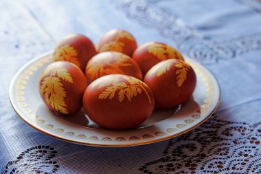 Görüntü, sarı yaprak desenli yedi kahverengi yumurtayı altın kenarlı beyaz bir tabakta, mavi ve beyaz dantel bir masa örtüsüne yerleştirilmiş olarak gösteriyor..