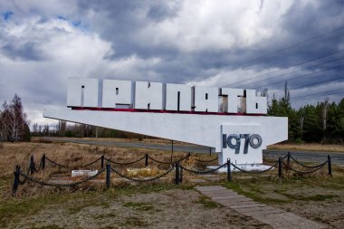 Çorak bir araziyle çevrili büyük beyaz bir tabela. Kiril alfabesi ile metin: Pripyat, şehrin adı.