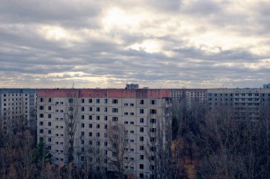 Bulutlu gökyüzü, uzun ve çorak ağaçların arasına kurulmuş eski apartman binalarının üzerinde beliriyor..