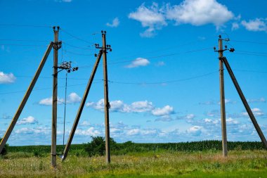 Elektrik direkleri, parçalı bulutlu bir gökyüzünün altında birbirine bağlı tellerle çimenli bir alanda duruyor..