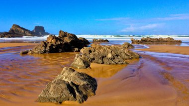 Sahil manzarası sakin bir sahil, dağınık kayalar ve parlak bir gökyüzünün altındaki uzak kayalıklar..