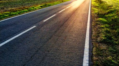 Güneş boş bir yolda beyaz kenarlı çimlerin arasında parlıyor..