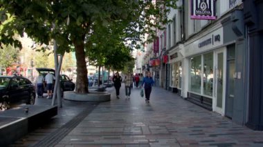 Cork, İrlanda, 30 Eylül 2022. Yayalar dükkanların ve işyerlerinin önünden geçerken, ağaçlarla çevrili bir şehir caddesi hareketlilikle dolup taşıyor, havada normallik hissi var. Mantar halkı..