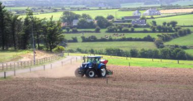 County Cork, İrlanda, 20 Ağustos 2022. Bir traktör toprakta çalışır, toz bulutlu gökyüzü ve yuvarlanan arazi zemininde yükselir. Bir mavi traktör yazın ekili bir tarlayı işliyor ve ekiyor..