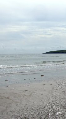 Küçük dalgaların kıyıya vurduğu kumlu bir sahil. Su berraktır ve güneş ışığını yansıtır. Işıldayan bir etki yaratır. Gökyüzü bulutlu ve mavi bir bakış var..