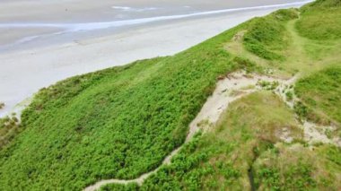 Çimenli bir kumulun havadan görünüşü ve bitkilerin arasından geçen bir patika. Arka planda kumlu bir sahil ve okyanus görülebilir. Önünde çimenli bir tepe olan kumlu bir sahil..