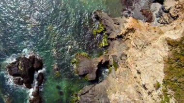 Sahne, arka planda sakin bir su kütlesiyle kayalık bir kıyı şeridini tasvir ediyor. Su sakin ve şeffaf. Dalgalar dağılmış kayalara çarpıyor..