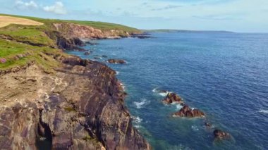 Kıyı şeridi kayalık ve arkasında çimen kaplı bir yamaç var. Deniz sakin ve gökyüzü bulutsuz..