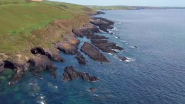 Üzerinde yeşil bir tepe olan kayalık bir uçurum manzarası. Uçurumun önünde berrak mavi bir deniz ve üzerinde bulutlar olan mavi bir gökyüzü var..