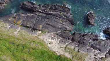 Bir dizi koyu gri kaya kısmen berrak, turkuaz suya batar. Kayalar beyaz köpüklü dalgalarla çevrili..