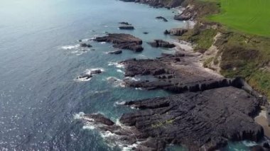 Yüksek bir perspektiften bakınca kıyı manzarası. Odak noktası mavi okyanusa doğru uzanan kayalık bir çıkıntı. Kayalar koyu gri ve pürüzlüyken su berrak ve turkuazdır..